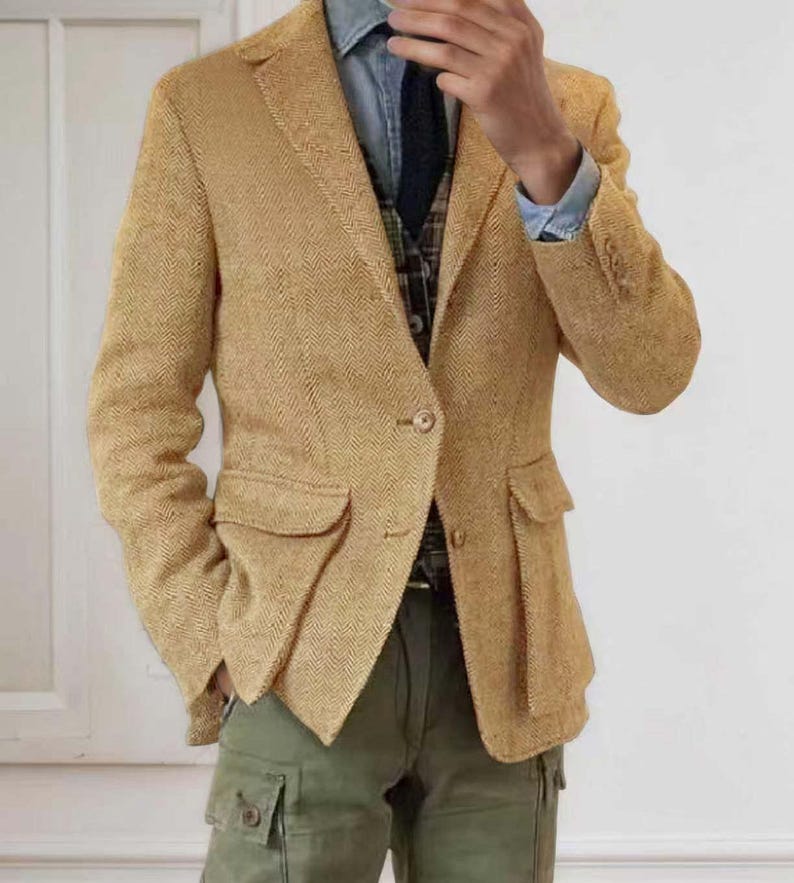 May include: A tan herringbone blazer with two front pockets and a single-button closure. The blazer is paired with a denim shirt, a patterned vest, a dark tie, and olive green cargo pants. The outfit is a blend of textures and colors.