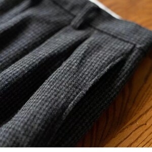 K&ouml;nnte beinhalten: Nahaufnahme eines dunkelgrauen Gewebes mit einem dezenten Hahnentrittmuster. Der Stoff ist dick und scheint eine Wollmischung zu sein, mit sichtbaren Falten und einem Taschen Detail. Die Textur deutet auf ein ma&szlig;geschneidertes Kleidungsst&uuml;ck hin.