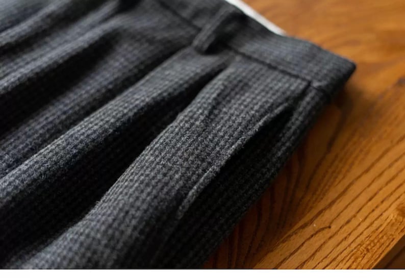 K&ouml;nnte beinhalten: Nahaufnahme eines dunkelgrauen Gewebes mit einem dezenten Hahnentrittmuster. Der Stoff ist dick und scheint eine Wollmischung zu sein, mit sichtbaren Falten und einem Taschen Detail. Die Textur deutet auf ein ma&szlig;geschneidertes Kleidungsst&uuml;ck hin.