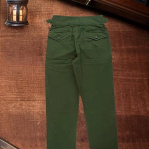 Puede incluir: Pantalones de color verde oliva con dise&ntilde;o de cintura alta, pesta&ntilde;as de ajuste laterales, dos bolsillos traseros con solapas abotonadas y corte de pierna recta. Los pantalones presentan una cinturilla estructurada y costuras visibles.
