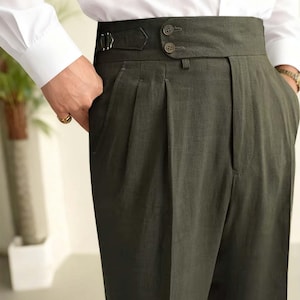 May include: Dark green high-waisted trousers with a wide waistband and pleats. The trousers have two buttons and a buckle detail. The person is wearing a white dress shirt and a gold ring.