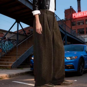 May include: High-waisted, wide-leg trousers in a dark olive-brown textured fabric. These full-length pants feature a pleated front design, a structured waistband with belt loops, and a loose, flowing silhouette that extends down to the ankles.