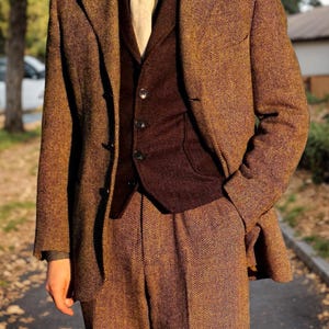 May include: A three-piece brown herringbone tweed suit. The suit includes a blazer, vest, and trousers. The blazer has a notched lapel and button closure. The vest is a darker brown color. The suit is paired with a light-colored tie and a patterned shirt.