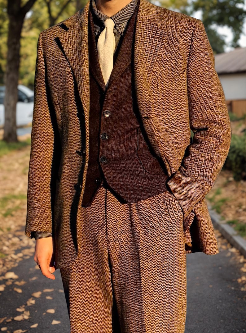 May include: A three-piece brown herringbone tweed suit. The suit includes a blazer, vest, and trousers. The blazer has a notched lapel and button closure. The vest is a darker brown color. The suit is paired with a light-colored tie and a patterned shirt.