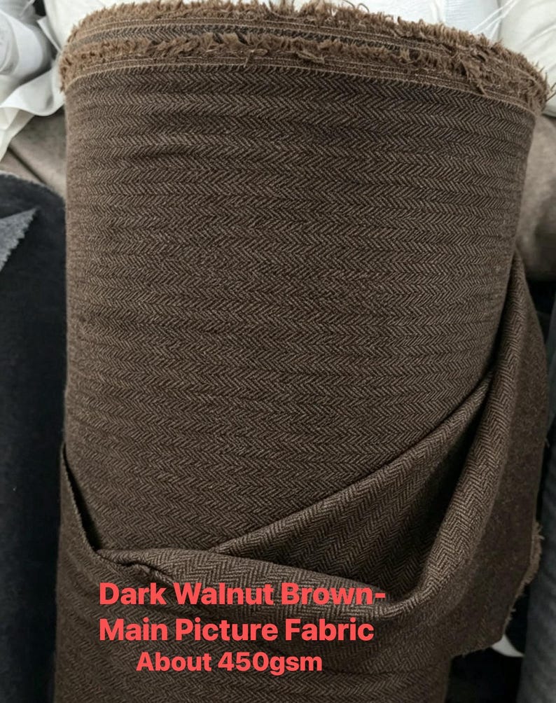 May include: A roll of dark walnut brown fabric featuring a woven herringbone pattern. The textile has a visible frayed edge at the top and a weight of about 450gsm as indicated by the text printed on the lower portion of the fabric roll.
