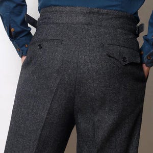 May include: Dark gray trousers featuring a classic herringbone pattern, a high-waisted design with side adjustment tabs, and two back pockets with buttoned flaps. The trousers are constructed from a textured fabric and include a visible center seam.