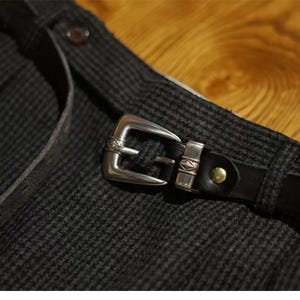 K&ouml;nnte beinhalten: Nahaufnahme eines dunkelgrauen Stoffes mit Muster, einem schwarzen Lederg&uuml;rtel und einer silbernen Schnalle. Die Schnalle ist rechteckig mit dekorativen Details. Ein kleiner, runder Knopf ist auf dem Stoff sichtbar.