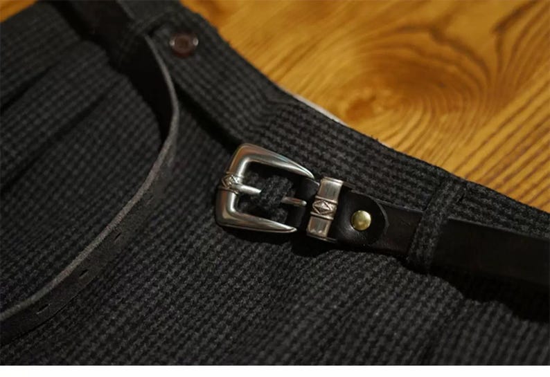 K&ouml;nnte beinhalten: Nahaufnahme eines dunkelgrauen Stoffes mit Muster, einem schwarzen Lederg&uuml;rtel und einer silbernen Schnalle. Die Schnalle ist rechteckig mit dekorativen Details. Ein kleiner, runder Knopf ist auf dem Stoff sichtbar.