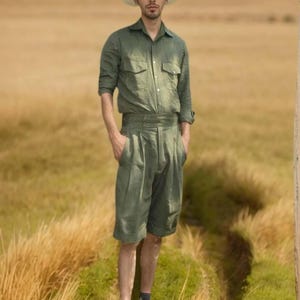 May include: A person wearing a light green button-down shirt with two chest pockets, matching shorts, and a straw hat. The outfit is paired with white shoes and dark socks. The background is a field of tall grass.