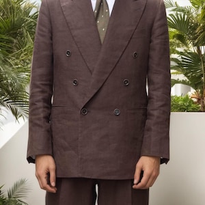 Puede incluir: Una americana cruzada de color marrón oscuro con solapas de pico anchas y seis botones, sobre una camisa blanca de cuello y una corbata verde oliva oscuro con pequeños lunares blancos y rojos, combinada con pantalones marrón oscuro.