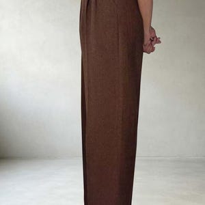 May include: High-waisted brown trousers featuring a straight-leg cut, a thin brown leather belt at the waist, and cuffed hems. The trousers are paired with dark brown leather loafers, showcasing a clean, structured silhouette from a side profile.