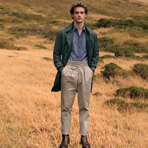 May include: A person wearing a dark green overcoat, a denim shirt, and khaki pants with brown leather boots. The pants have a high waist and are cuffed at the ankles. The background is a field of dry grass.