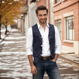 Könnte beinhalten: Eine dunkelblaue Weste mit drei hellen Knöpfen über einem weißen langärmeligen Hemd. Das Outfit wird mit einer dunkelblauen Jeans, einem braunen Ledergürtel mit Metallschnalle und einer silbernen Armbanduhr kombiniert.