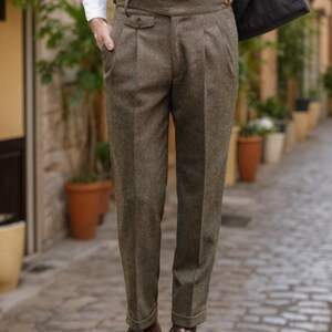 May include: High-waisted brown trousers featuring a textured wool-like fabric, double pleats at the front, a wide waistband with a side buckle adjuster, and cuffed hems. The trousers are paired with a white button-down shirt and brown leather shoes.