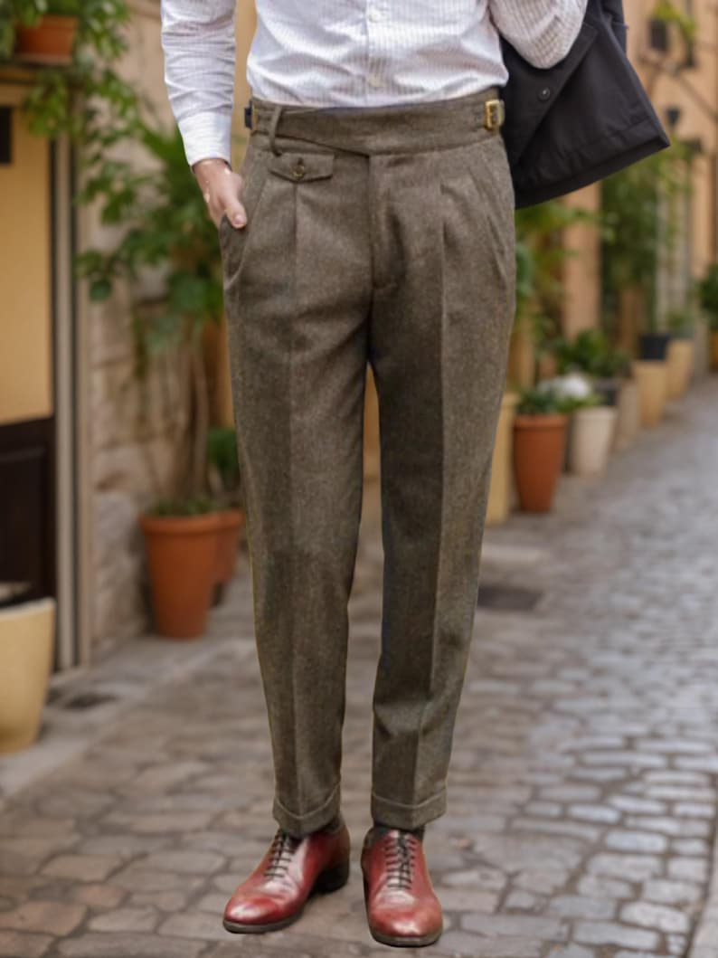 May include: High-waisted brown trousers featuring a textured wool-like fabric, double pleats at the front, a wide waistband with a side buckle adjuster, and cuffed hems. The trousers are paired with a white button-down shirt and brown leather shoes.