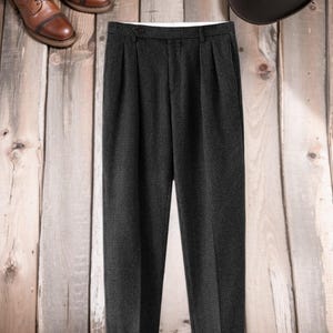 K&ouml;nnte beinhalten: Dunkelgraue, plissierte Hose mit dezentem Karomuster. Die Hose wird mit braunen Lederstiefeln und einem dunkelbraunen Hut auf einem Holzgrund gezeigt. Die Hose hat einen Knopfverschluss und einen wei&szlig;en Innenbund.