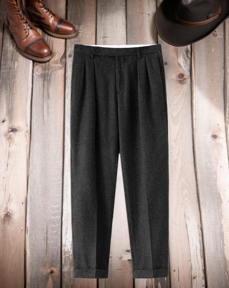 K&ouml;nnte beinhalten: Dunkelgraue, plissierte Hose mit dezentem Karomuster. Die Hose wird mit braunen Lederstiefeln und einem dunkelbraunen Hut auf einem Holzgrund gezeigt. Die Hose hat einen Knopfverschluss und einen wei&szlig;en Innenbund.