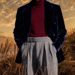 Puede incluir: Un blazer de terciopelo azul marino con diseño de doble botonadura, combinado con un cuello alto burdeos y pantalones de espiga grises. El conjunto se presenta sobre un fondo de hierba alta y cielo de atardecer.