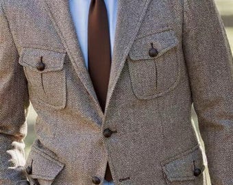 Puede incluir: Chaqueta de tweed marrón con diseño de espiga, cuatro bolsillos delanteros y botones marrones oscuros. La chaqueta se combina con una camisa azul claro y una corbata marrón. Corte clásico y entallado.