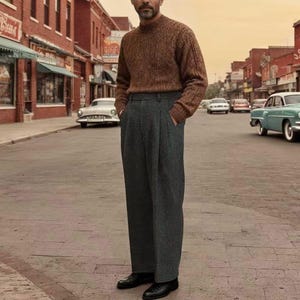 Op de afbeelding: Een persoon in een bruine kabelgebreide trui en een grijze plooibroek staat op een straat met bakstenen. Hij draagt zwarte leren schoenen. Klassieke auto's staan langs de straat geparkeerd.