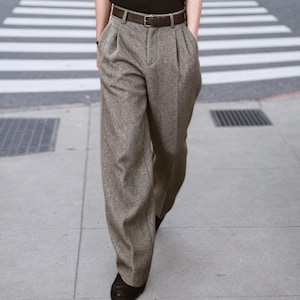 May include: High-waisted trousers in a textured light brown and grey speckled fabric. These wide-leg pants feature front pleats, a center zipper fly, and are secured at the waist with a dark brown leather belt featuring a silver-toned metal buckle.