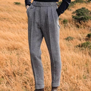 May include: Grey herringbone tweed trousers with a pleated front and cuffed hem. The tailored trousers feature side pockets. The fabric has a subtle pattern, offering a classic and versatile look.