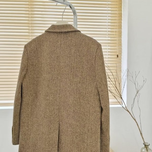 May include: A brown tweed blazer hangs on a metal rack. The blazer has a classic notched lapel and a two-button closure. The fabric has a subtle herringbone pattern. The blazer is a neutral color, suitable for various outfits.