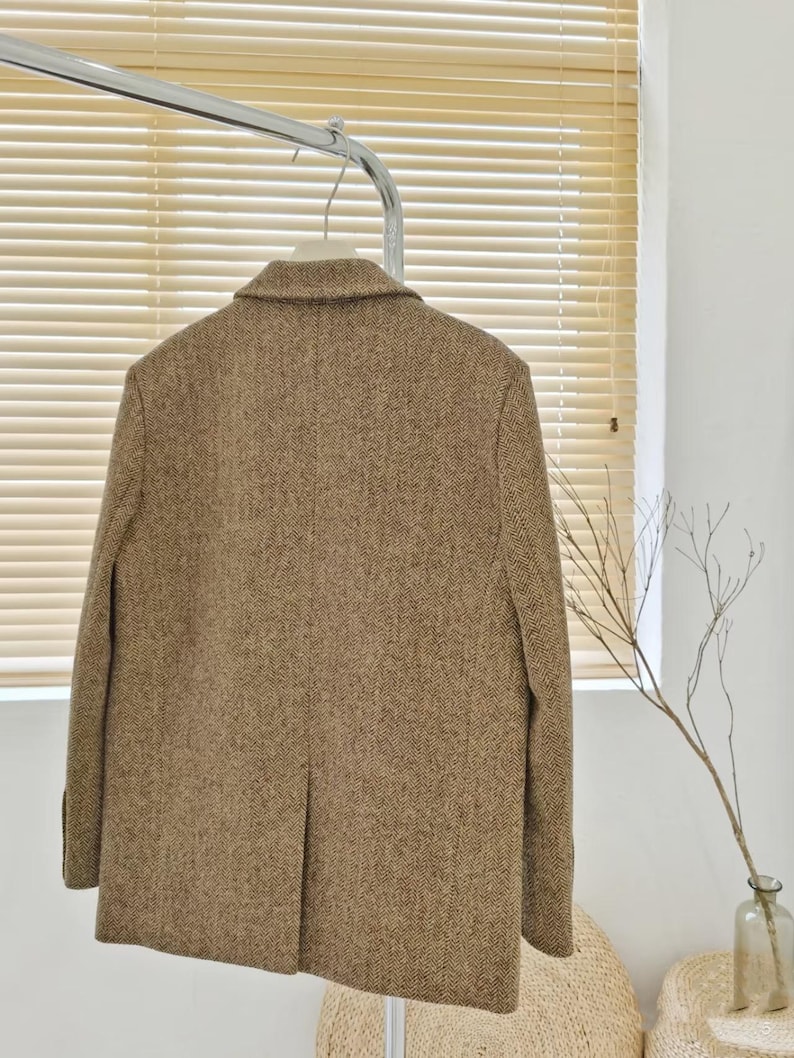May include: A brown tweed blazer hangs on a metal rack. The blazer has a classic notched lapel and a two-button closure. The fabric has a subtle herringbone pattern. The blazer is a neutral color, suitable for various outfits.