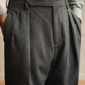 K&ouml;nnte beinhalten: Nahaufnahme einer grauen Hose mit Hahnentrittmuster und Falten. Die Hose hat einen Knopfverschluss und G&uuml;rtelschlaufen. Der Stoff scheint eine Wollmischung zu sein. Die Hose ist Teil einer klassischen Herrenkollektion.