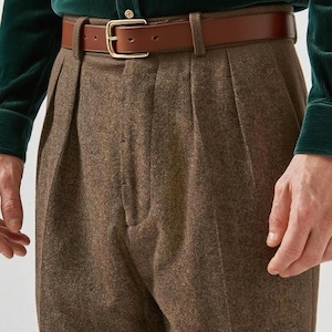 May include: High-waisted brown wool trousers featuring double pleats at the front, paired with a smooth tan leather belt that has a gold-toned metal buckle. The trousers are worn with a dark green corduroy long-sleeve shirt with buttons.