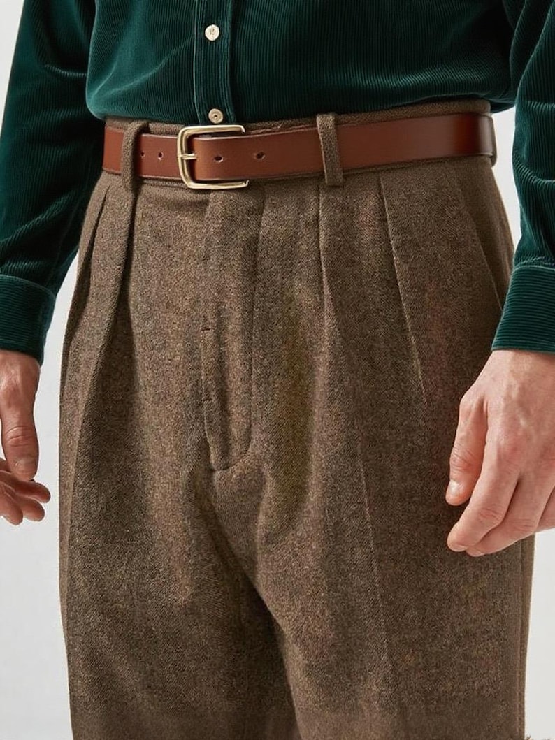 May include: High-waisted brown wool trousers featuring double pleats at the front, paired with a smooth tan leather belt that has a gold-toned metal buckle. The trousers are worn with a dark green corduroy long-sleeve shirt with buttons.