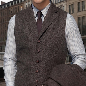 May include: A brown herringbone waistcoat with matching buttons and a notched lapel. The waistcoat is worn over a striped shirt and a dark tie. A matching jacket is held in the arm.