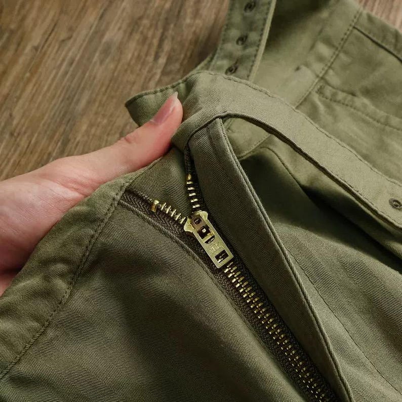 Puede incluir: Pantalones de color verde oliva con una cremallera met&aacute;lica resistente y un tirador de tono lat&oacute;n. La tela muestra un patr&oacute;n de espiga distintivo y la prenda incluye una cintura reforzada con detalles de costuras visibles.