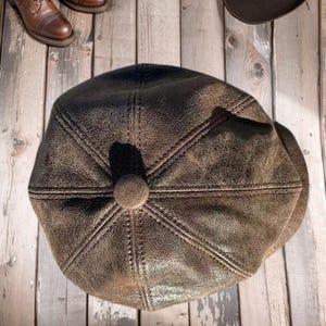 Puede incluir: Gorra de estilo newsboy de cuero marrón con un botón en la parte superior. La gorra tiene un aspecto envejecido y texturizado, con costuras visibles. También se ven botas de cuero marrón y un sombrero marrón oscuro.