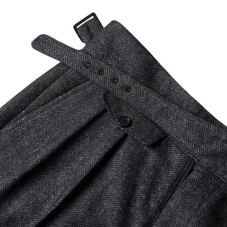May include: A pair of dark gray trousers featuring a classic herringbone pattern. The waistband includes an adjustable side strap with a metal buckle and a small triangular tab secured by a single black button, along with visible vertical pleats.