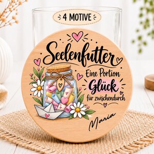 Könnte beinhalten: Ein runder Holzdeckel mit einem Aufdruck, der ein Glas mit bunten Papierrollen, einem Papierboot und Gänseblümchen zeigt. Der Text lautet Seelenfutter, Eine Portion Glück für zwischendurch sowie der Name Maria, mit 4 MOTIVE oben.