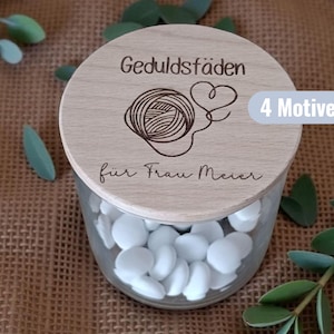 Könnte beinhalten: Ein klares Glas mit weißen Bonbons, bedeckt mit einem Holzdeckel. Der Deckel ist mit den Worten "Geduldsfäden" und einem Garnknäuel-Design graviert. Der Text "4 Motive" befindet sich auf einem blauen Etikett.