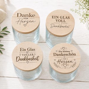 Könnte beinhalten: Vier klare Gläser mit Holzdeckeln. Die Deckel tragen deutsche Texte und Herzmotive. Die Gläser stehen auf einer hellen Holzoberfläche, im Hintergrund ein weißes Tuch und Grünzeug. Auf den Deckeln stehen Sätze wie "Danke von Herzen" und "Ein Glas voller Dankbarkeit."
