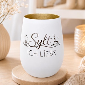 Könnte beinhalten: Weißes, stieloses Weinglas mit goldenem Rand, mit dem Text "Sylt ICH LIEBS" und einer Grafik eines Leuchtturms und einer Insel. Das Glas steht auf einem Holzuntersetzer, im Hintergrund eine Vase mit Blumen und eine brennende Kerze.