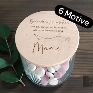 Könnte beinhalten: Ein Glas mit einem Holzdeckel, gefüllt mit weißen und rosa Bonbons. Der Deckel ist mit dem Namen "Marie" und einem deutschen Zitat graviert. Ein schwarzes Banner mit dem Text "6 Motive" befindet sich oben rechts. Ein Zweig mit grünen Blättern ist links.