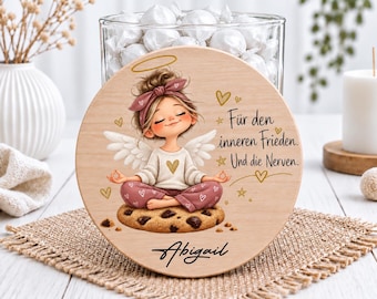 Könnte beinhalten: Ein rundes Holzschild mit einer Cartoon-Engel-Illustration mit Dutt und Stirnband, die im Lotussitz auf einem Schokoladenkeks sitzt. Der Text lautet: Für den inneren Frieden. Und die Nerven. Darunter ist der Name Abigail aufgedruckt.