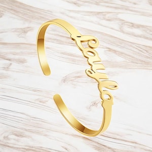 May include: A gold-coloured, open cuff bracelet with the name "Camila" in a cursive font. The bracelet is made of a shiny metal and rests on a light-coloured, wood-grain surface.