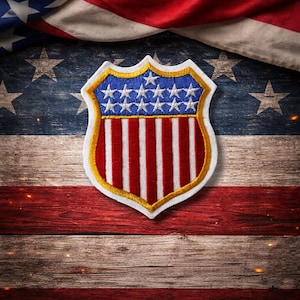 May include: A shield-shaped embroidered patch featuring a blue upper section with white stars and a lower section with vertical red and white stripes, outlined in gold and white thread, positioned against a rustic wooden American flag background.