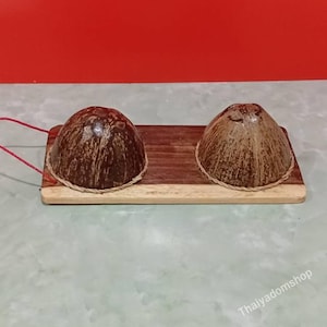 Può includere: Due metà di guscio di cocco testurizzate marrone scuro sono montate su una base rettangolare in legno. Ogni guscio è circondato da un sottile bordo in corda chiara. Un sottile filo rosso sporge dal lato sinistro della base in legno.