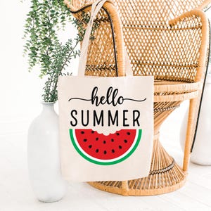 Peut inclure: Un sac fourre-tout en toile de couleur cr&egrave;me avec les mots "hello SUMMER" en &eacute;criture noire et en lettres majuscules. En dessous, une tranche de past&egrave;que rouge avec des graines noires et une peau verte. Le sac a des anses cr&egrave;me.