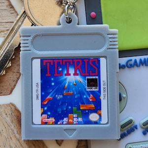 以下が含まれることがあります： 銀色のチェーンとキーリングが付いた、グレーのテトリスのゲームカートリッジキーホルダー。カートリッジには赤いテトリスのロゴと、ブロックが落ちる青い背景が描かれています。「MADE IN JAPAN」の文字が見えます。