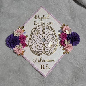 Op de afbeelding: Een versierde afstudeerhoed met de woorden "Psyched for the next Adventure B.S." in gouden letters. De hoed heeft een hersengrafiek, vlinders en bloemaccenten in roze, paars en goud.