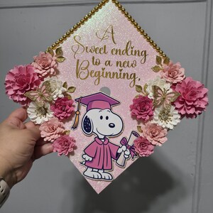 Puede incluir: Un gorro de graduaci&oacute;n rosa decorado con flores de papel, ribete dorado y un gr&aacute;fico de Snoopy con toga. El gorro tiene el texto "A sweet ending to a new beginning."