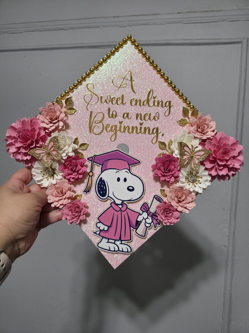 Puede incluir: Un gorro de graduaci&oacute;n rosa decorado con flores de papel, ribete dorado y un gr&aacute;fico de Snoopy con toga. El gorro tiene el texto "A sweet ending to a new beginning."