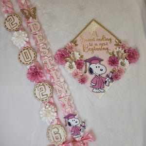 Puede incluir: Conjunto de lei y gorra de graduaci&oacute;n rosa y dorado. El lei deletrea el nombre "Destiny" e incluye el a&ntilde;o "2026". La gorra presenta a Snoopy con toga de graduaci&oacute;n y el texto "A Sweet ending to a new Beginning."